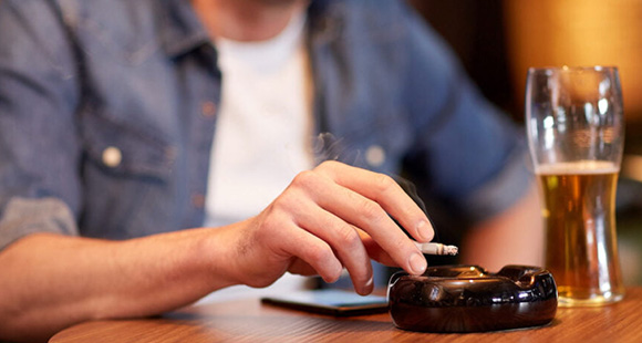 a person sat in a pub with a pint of beer and smoking a cigarette in front of an ash tray