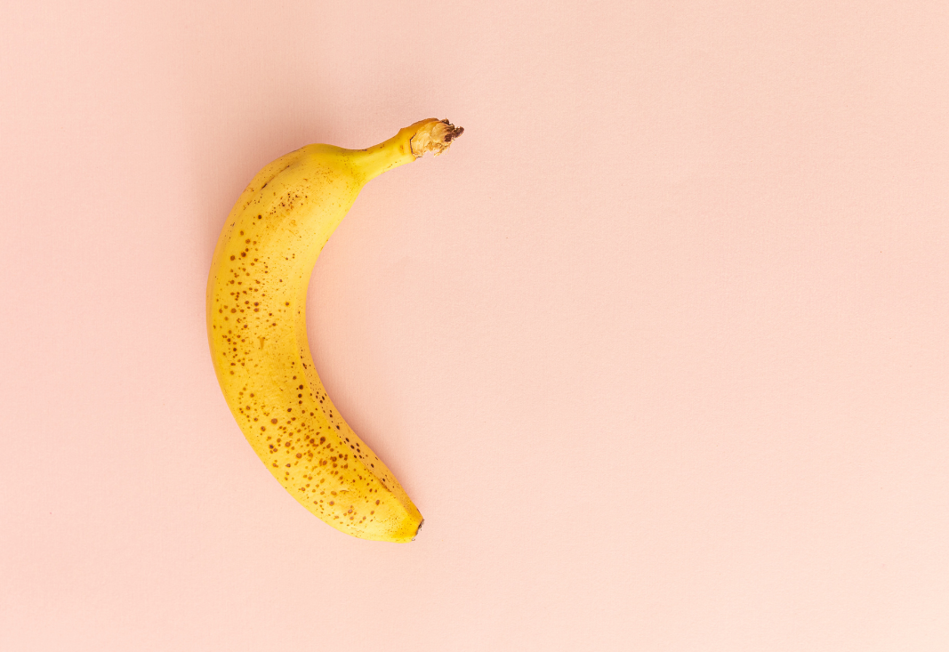 Single ripe banana with brown speckles on a light pink background, positioned to emphasise its curved shape with empty space around it.