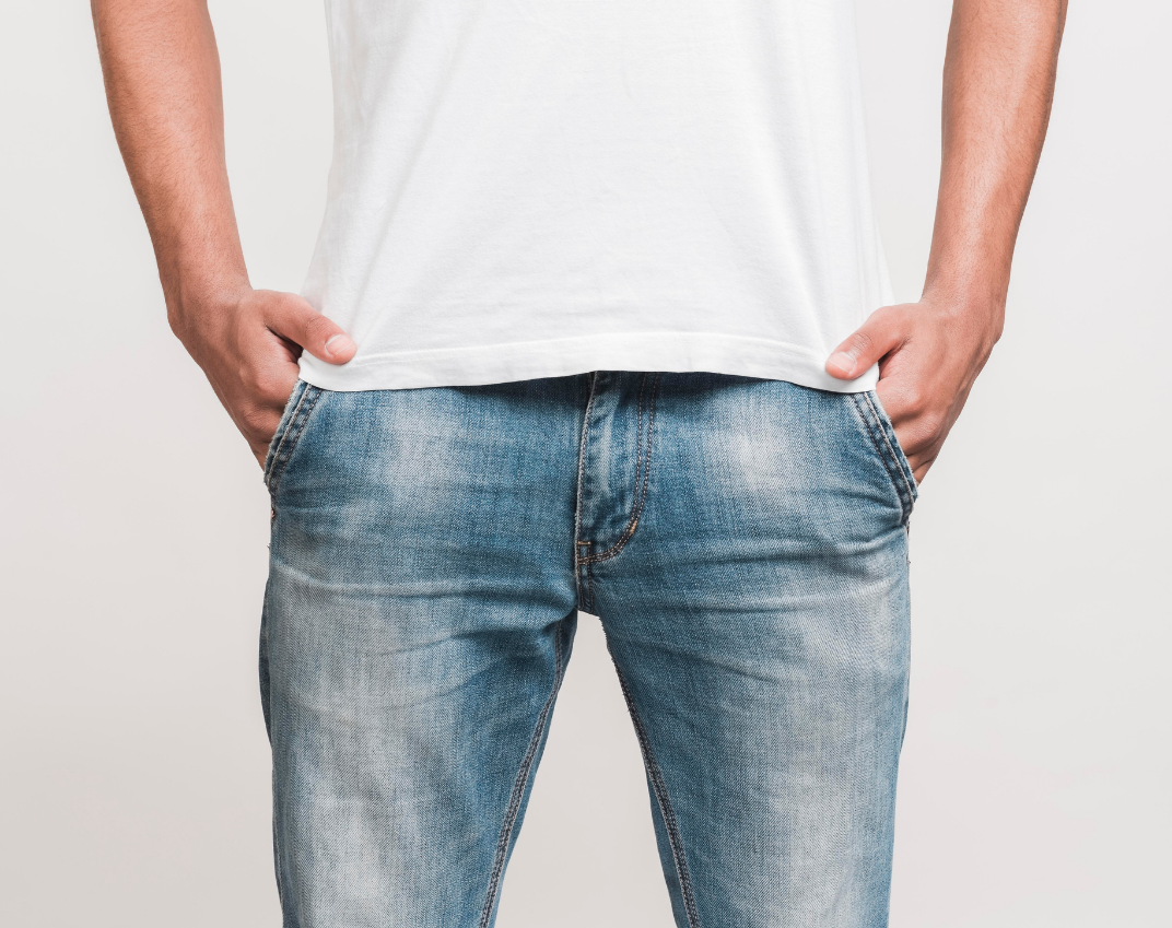 Close-up of a man wearing a white T-shirt and blue jeans, standing with both hands in his front pockets against a plain grey background.