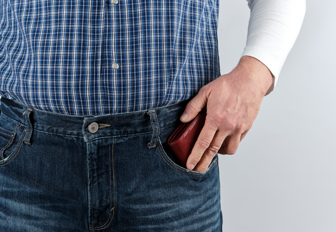 Close-up of a man wearing a blue checked shirt and jeans, placing a red wallet into his front trouser pocket.