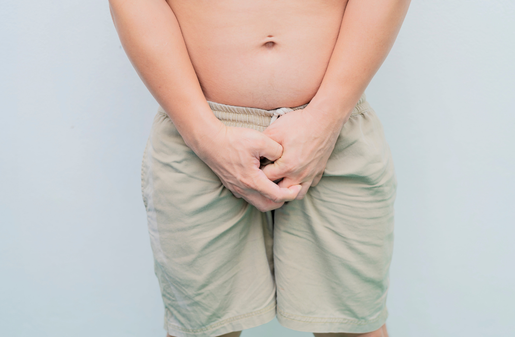Close-up of a shirtless man wearing light-coloured shorts, standing with both hands clasped over his groin area against a plain background.