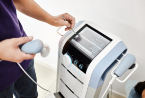 Healthcare professional operating a shockwave therapy machine, holding a treatment applicator and adjusting settings on a touchscreen control panel.