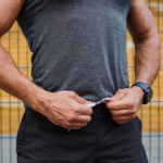 Close-up of a muscular man in a grey sleeveless athletic shirt and black shorts, standing outdoors and adjusting the waistband of his shorts with both hands. A fitness watch is visible on his wrist, with a metal fence blurred in the background.
