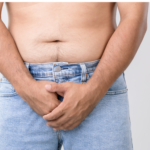 Close-up of a shirtless man wearing light blue jeans, standing with both hands clasped over his groin area against a plain grey background.