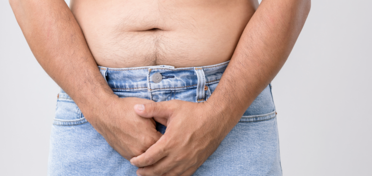 Close-up of a shirtless man wearing light blue jeans, standing with both hands clasped over his groin area against a plain grey background.
