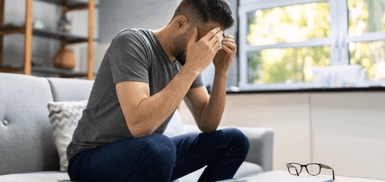 Man sitting on a sofa with his head in his hands, illustrating the emotional impact of leaving Peyronie’s disease untreated.