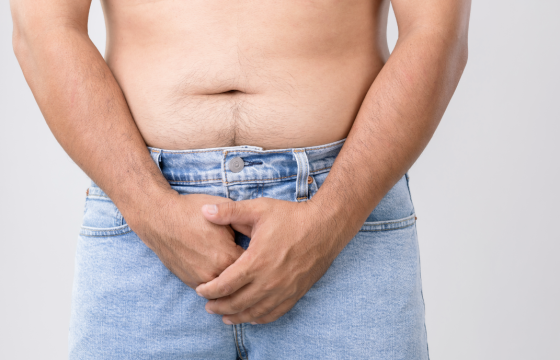 Close-up of a shirtless man wearing light blue jeans, standing with both hands clasped over his groin area against a plain grey background.